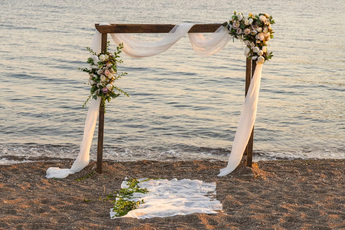 Wedding arch draped in white fabric and flowers on a quiet beach at golden hour
