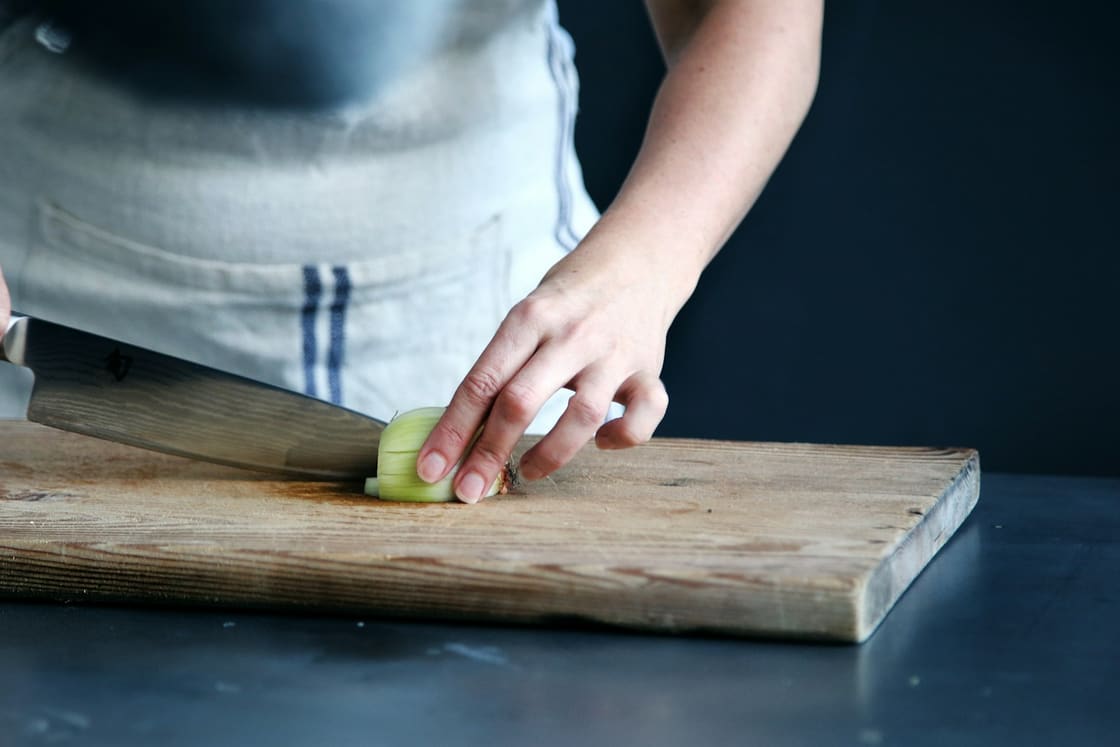 Cook in a linen apron slicing an onion with a chef’s knife on a wooden board