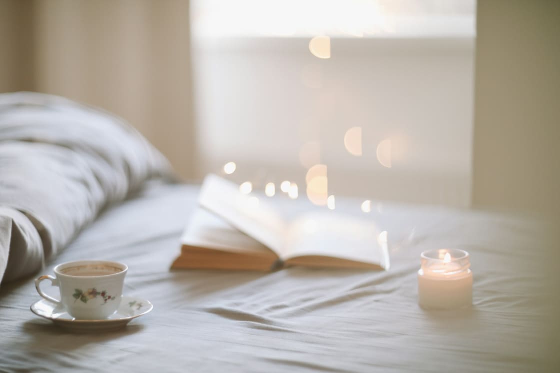 Soft-lit bed with a teacup, an open book and a lit candle on the covers