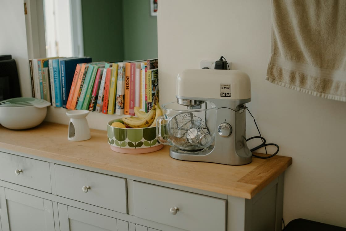 Cream stand mixer on a kitchen sideboard beside a fruit bowl and cookbooks